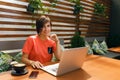 Portrait of confident mature professional woman in glasses, a coral T-shirt sitting on summer terrace in cafe, using laptop Royalty Free Stock Photo
