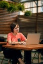 Portrait of confident mature professional woman in glasses, a coral T-shirt sitting on summer terrace in cafe, using laptop Royalty Free Stock Photo