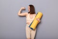Portrait of confident prod woman wearing beige top and leggins holding fitness mat over gray background, raised her arm,