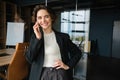 Portrait of confident saleswoman in suit, having a conversation on mobile phone, standing in her office near glass wall Royalty Free Stock Photo