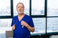 Portrait of confident senior adult male doctor in medical uniform standing in hospital office on background of window Royalty Free Stock Photo