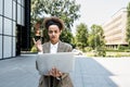 Portrait of confident successful young curly woman, formally dressed, business woman standing with laptop outdoors against the