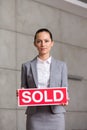 Portrait of confident young saleswoman holding sold placard while standing against wall in apartment