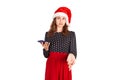 Portrait of a confused cute girl in dress pointing at mobile phone. emotional girl in santa claus christmas hat isolated on white Royalty Free Stock Photo