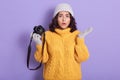 Portrait of confused helpless cute young female with black curly hair, raising one hand, holding photocamera, looking directly at