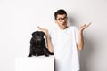 Portrait of confused hipster guy dog owner shrugging, standing near cute black pug pet, white background Royalty Free Stock Photo