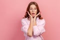 Portrait of confused young brunette woman in eyeglasses and hoodie touching face and looking with big eyes and open mouth, Royalty Free Stock Photo