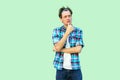 Portrait of confused young man in casual blue checkered shirt and headband standing, touching chin, thinking and concentrated to Royalty Free Stock Photo