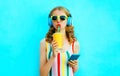 Portrait cool girl drinking fruit juice holding phone listening to music in wireless headphones on colorful blue