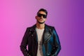 Portrait of cool serious young Arab man in sunglasses wearing black leather jacket, looking at camera in neon light