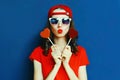 Portrait cool young woman with heart shaped red lollipop blowing red lips sending sweet air kiss wearing baseball cap, sunglasses Royalty Free Stock Photo