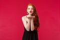 Portrait of coquettish feminine young beautiful redhead woman in black stylish dress, sending air kiss on palms near Royalty Free Stock Photo