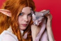 Portrait of cosplay elf young redhead woman with Sphynx kitten on red background Royalty Free Stock Photo