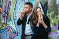 portrait of couple in luxury clothing eating hot dogs near wall with graffiti