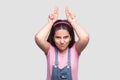 Portrait of crazy brunette young girl in casual pink t-shirt and blue overalls standing and looking at camera with cow horns