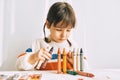 Portrait of creative cute little girl playing with oil pencils, sitting at white desk at home. Pretty preschool kid draws Royalty Free Stock Photo