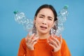 Portrait of crying asian woman with two plastic bottles, tired of recycling, sorting rubbish, blue background