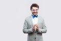 Portrait of cunning handsome bearded man in casual grey suit and blue bow tie standing with funny face and palm hand looking at Royalty Free Stock Photo