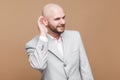 Portrait of cunning handsome middle aged bald bearded businessman in classic light gray suit standing and holding hand on ear and