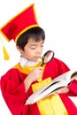 Portrait Of Curious Boy In Red Gown Kid Graduation With Mortarboard Looking A Book Through Magnifying Glass Royalty Free Stock Photo