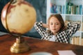 Portrait of curious child girl sitting on chair with hands behind head in front of table with a small globe and looking Royalty Free Stock Photo