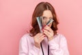 Portrait of curious curly haired teenage girl in hoodie peeping through bunch of dollar banknotes