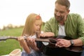 Portrait of curious little girl sitting with her loving father on a green grass in park and learning how to play guitar Royalty Free Stock Photo