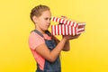 Portrait of curious nosy little girl with braid in denim overalls peeking into gift box, in anticipation of surprise