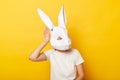 Portrait of curious woman wearing white T-shirt and paper rabbit mask standing isolated over yellow background, keeps hand near Royalty Free Stock Photo