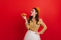 Portrait of curly French girl eating crispy croissant on red background. Dark-haired woman in checkered blouse and Royalty Free Stock Photo