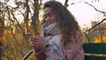 Portrait of curly-haired caucasian girl sitting on bench and watching gladly into smartphone in autumnal park.