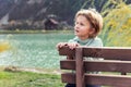 Portrait of cute adorable caucasian blond stylish 10 year old kid boy in hoodie sitting on wooden bench against pond at Royalty Free Stock Photo