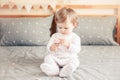 Caucasian blonde baby girl in white onesie sitting on bed in bedroom