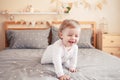 Caucasian blonde baby girl in white onesie sitting on bed in bedroom