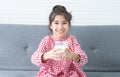 Portrait of cute Asian little girl smiling, holding and drinking a glass of milk, sitting on sofa at home, looking at camera. Royalty Free Stock Photo