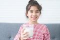 Portrait of cute Asian little girl smiling, holding and drinking a glass of milk, sitting on sofa at home, looking at camera. Royalty Free Stock Photo