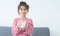 Portrait of cute Asian little girl smiling, holding and drinking a glass of milk, sitting on sofa at home, looking at camera. Royalty Free Stock Photo