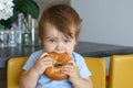 Portrait of cute baby boy with stylish haircut holding and eating big bagel sitting on yellow chair at home kitchen Royalty Free Stock Photo