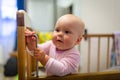 Portrait of cute baby girl with blue eyes is standing in crib. Adorable infant is standing up and smiling. Concept Royalty Free Stock Photo