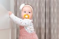 Portrait of cute baby girl in a dress, with a pacifier in her mouth and with a headband on her head, stands near the crib. The Royalty Free Stock Photo