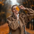 Portrait of a cute beautiful blonde woman in elegant beige coat in a vintage white sweater with a maple orange leaf near face Royalty Free Stock Photo