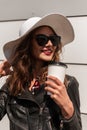 Portrait cute beautiful smiling woman with red lipstick in hat in black leather jacket in sunglasses with cup coffee near modern Royalty Free Stock Photo