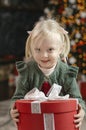 Portrait of cute blonde three-year-old girl with large gift on Christmas tree and garlands background