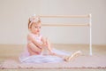 Portrait of cute blue-eyed baby ballerina in pink sitting near the ballet barre putting on pointe shoes Royalty Free Stock Photo