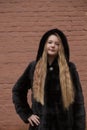 Portrait of cute brunette teen girl in black fur coat on the background of brown brick wall . Cosiness, fashion, style Royalty Free Stock Photo