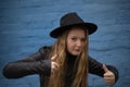 Portrait of cute brunette teen girl in Leather Jacket and black hat showing big thumbs up, on the background of blue brick wall . Royalty Free Stock Photo