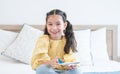 Portrait of cute Caucasian little girl with pony tails hair sitting on bed at home and playing with airplane model toy, holding Royalty Free Stock Photo