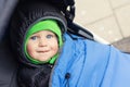 Portrait of cute caucasian toddler boy in black hooded warm insulated dawn overall jacket and blue blanket sitting in stroller and Royalty Free Stock Photo