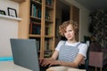 Portrait of cute curly little school girl sitting at table looking at camera doing homework using laptop and paper Royalty Free Stock Photo