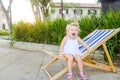 Portrait of cute emotional blondy toddler girl in white dress sitting on the deckchair and yelling. City park recreation area. Sel Royalty Free Stock Photo